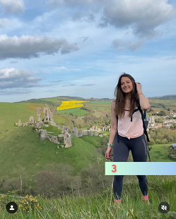 A white female with long brown hair smiles at the camera on a hiking path. Corfe Castle is in the background